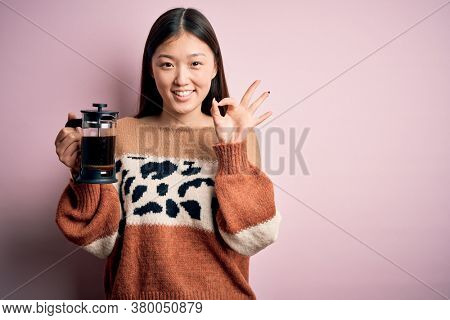 Young asian woman making a glass of coffe using french press coffee maker over pink background doing ok sign with fingers, excellent symbol