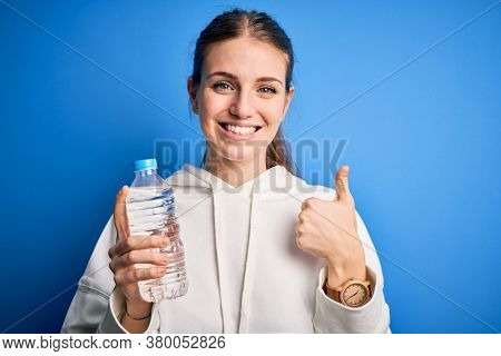 Young beautiful redhead woman doing sport drinking bottle of water over blue background happy with big smile doing ok sign, thumb up with fingers, excellent sign