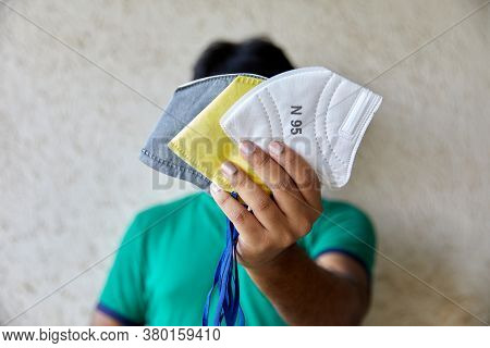 Man With Black Hair Holds Up Three Colourful Masks In Natural Light In Front Of Camera