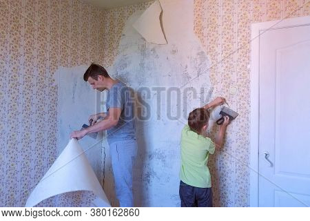 Father And Son Making Renovation Removing Wallpapers From Wall Together Using Spatulas. Construction