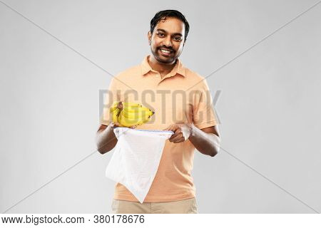 food, sustainability and eco living concept - happy smiling young indian man in polo t-shirt putting bananas into reusable string bag over grey background