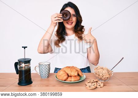 Young beautiful brunette woman eating breakfast holding cholate donut smiling happy and positive, thumb up doing excellent and approval sign 
