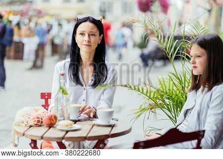Mom And Her Teenager Daughter Drink Coffee, Stiitng At Outdoor Cafe And Trying Socializing.