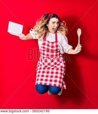 Young beautiful blonde cook woman wearing apron smiling happy. Jumping with smile on face holding recipe book and spoon over isolated red background