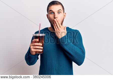 Young handsome man dirnking cola refreshment beverage covering mouth with hand, shocked and afraid for mistake. surprised expression 