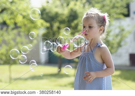Cute Little Girl Is Blowing Soap Bubbles With Pink Bubble Wand Outdoors.