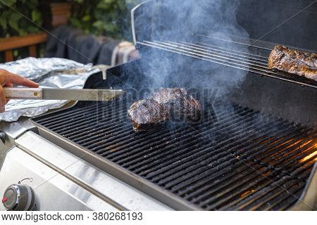 Man Cooking Beef On The Grill Outside, Flipping The Meat With Tongs.