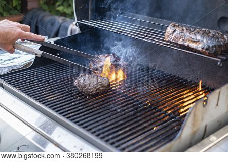 Man Cooking Beef On The Grill Outside, Flipping The Meat With Tongs.
