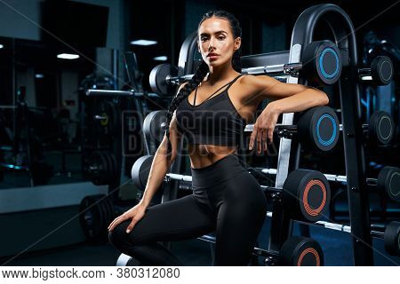 Side Close Up Of Female Bodybuilder With Long Braids Posing Near Stand With Sports Equipment. Srtong