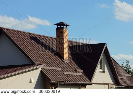 Brown Tiled Roof Of A Gray Private House With A Window And One Brick Chimney Against A Blue Sky