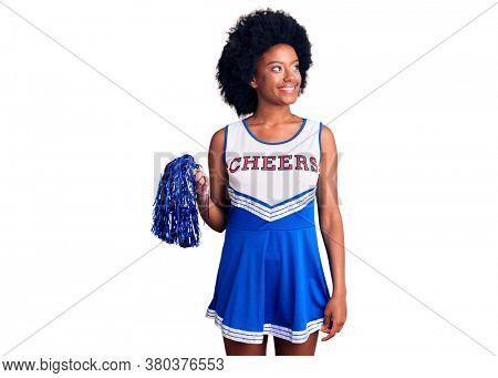 Young african american woman wearing cheerleader uniform holding pompom looking away to side with smile on face, natural expression. laughing confident. 