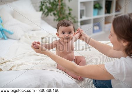 Cute Baby Sitting With Support On Bed. Mother Supports Child Sitting, Spreading Her Hands To Side. C