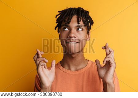 Smiling african manstanding against yellow background with crossed fingers and looking up, having pleading expression asking for good luck