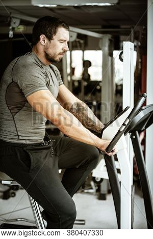 Fitness in gym, sport and healthy lifestyle concept. Handsome athletic man in grey shirt trains on velosimulator Bodybuilder male model with trained body does cardio workout.