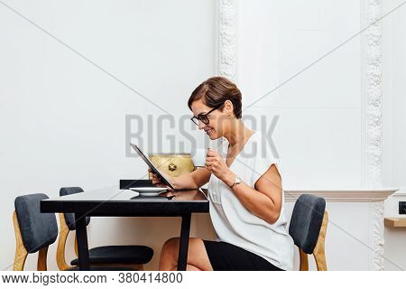 Side View Of A Woman Sitting At Table And Reading From Digital Tablet At Apartment. Businesswoman Ho