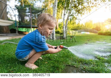 Cute Adorable Caucasian Blond Toddler Boy Enjoy Having Fun Watering Garden Flower And Lawn With Hose