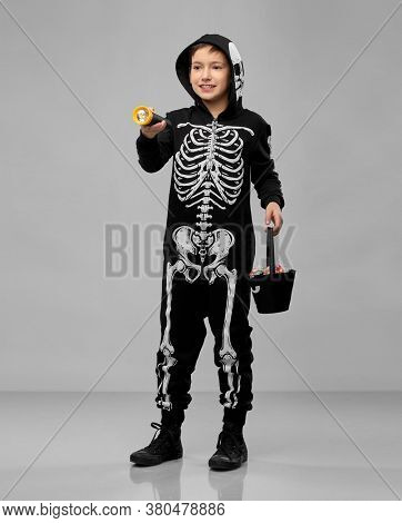 halloween, holiday and childhood concept - boy in black costume of skeleton with bucket of candies and flashlight trick-or-treating over grey background