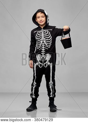 halloween, holiday and childhood concept - smiling boy in black costume of skeleton with candies trick-or-treating over grey background