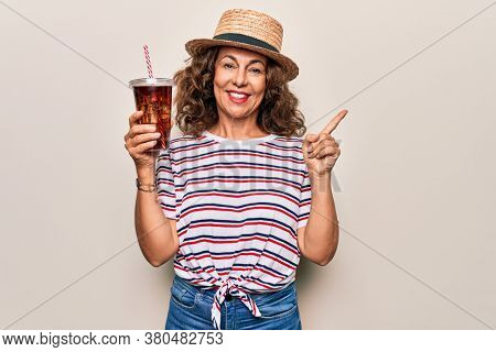 Middle age brunette woman on summer hat drinking fresh cola refeshment over isolated background smiling happy pointing with hand and finger to the side