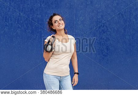 Young Women Keeps Headphones In Hand And Stays Near A Blue Wall