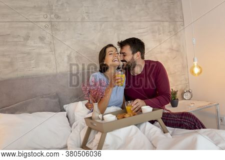 Beautiful Young Couple In Love Wearing Pajamas Sitting In Bed, Man Surprising Woman By Bringing Her 