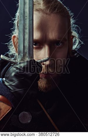 Close-up portrait of a medieval knight in armour and with a sword over black background. Historical reenactment.