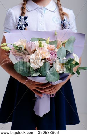 A Bouquet Of Flowers, Packed In Purple Paper In The Hands Of A Girl With Pigtails On A Light Backgro
