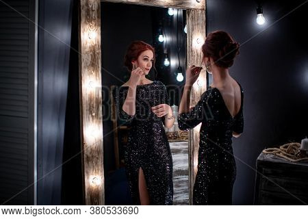 Beautiful, Elegant Woman In A Shiny Black Evening Dress Stands Next To A Dark High Mirror With Make-