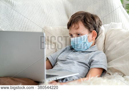 Focused Boy In A Mask, Lying On The Couch With A Laptop During The Quarantine Period. He Does Not Go