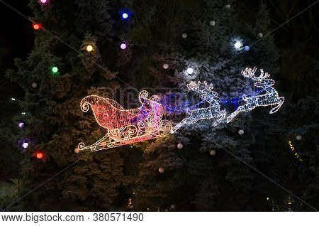 Flying Sleigh Of Santa Claus Against The Background Of Large Trees. Christmas Decoration From Led La