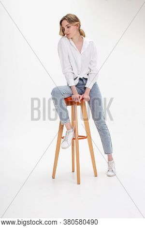 Portrait Of Young Pensive Caucasian Woman Posing In Shirt And Blue Jeans, Sitting On Stool In White 