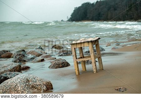 An Old Wooden Chair Standing Alone On The Shore Of A Raging Sea At Sunset In Gloomy Stormy Weather.