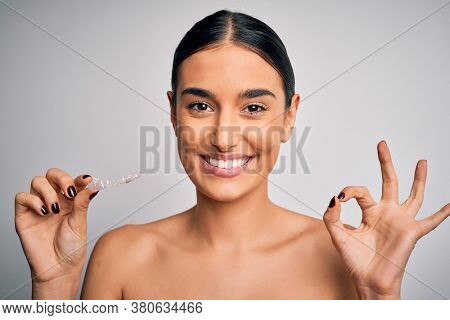 Young beautiful brunette woman holding dental aligner orthodontic to teeth corretion doing ok sign with fingers, excellent symbol