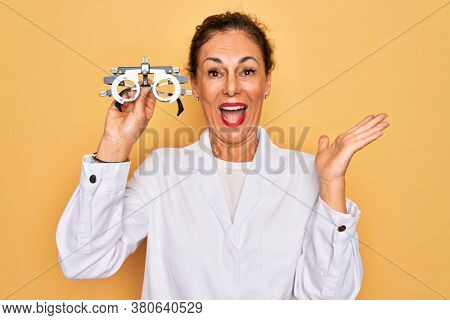 Middle age senior oculist woman holding optometrical glasses for vision correction very happy and excited, winner expression celebrating victory screaming with big smile and raised hands