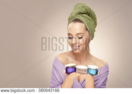 Happy Woman With Clean Fresh Skin Hold A Different Of Face Care Creams, Wearing In Bathrobe And Towe