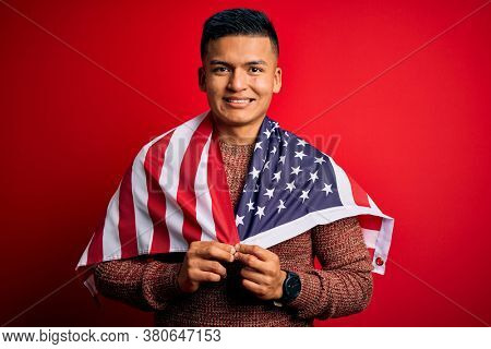 Young handsome latin patriotic man wearing united states of america falg with a happy face standing and smiling with a confident smile showing teeth