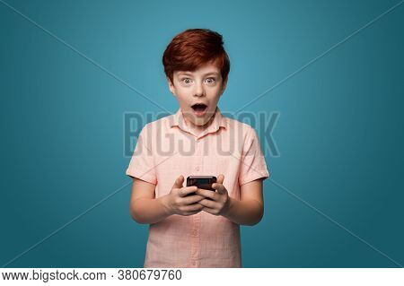 Shocked Caucasina Ginger Boy Holding A Phone And Looking At Camera On A Blue Studio Wall