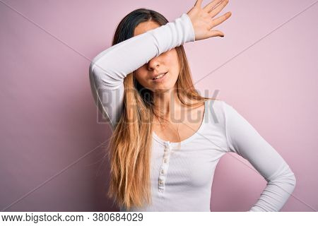 Young beautiful blonde woman with blue eyes wearing white t-shirt over pink background covering eyes with arm, looking serious and sad. Sightless, hiding and rejection concept