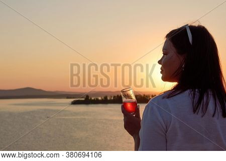 A Plump Girl Stands On The Shore Of A Lake With A Glass Of Wine At Sunset