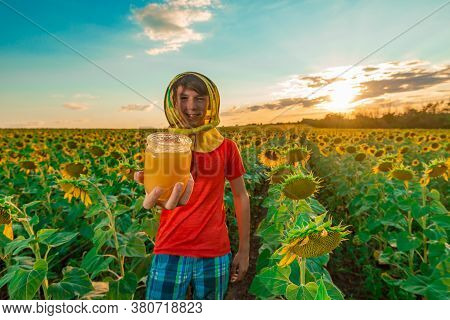 A Jar Of Honey On A Background Of Sunflower. A Boy Wearing A Mask From Bees Holds Out In His Hand A 