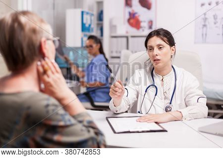 Senior Woman Having Problem With Sore Neck. Young Doctor Wearing White Coat And Stethoscope During E