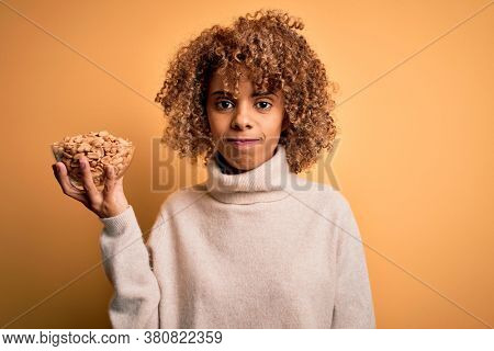 Young african american woman with curly hair holding bowl with healthy peanuts with a confident expression on smart face thinking serious