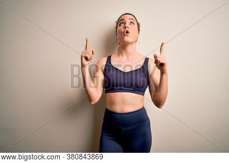 Young beautiful blonde sportswoman doing sport wearing sportswear over white background amazed and surprised looking up and pointing with fingers and raised arms.
