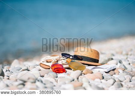 Beach Hat On Opened Book With Sunscreen And Shoes On Pebble Beach