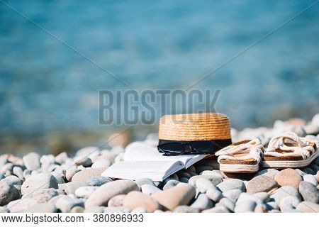 Beach Hat On Opened Book With Sunscreen And Shoes On Pebble Beach