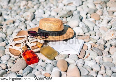 Beach Hat On Opened Book With Sunscreen And Shoes On Pebble Beach