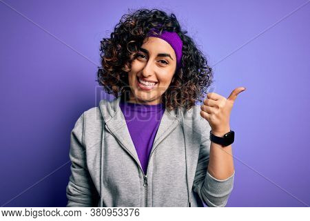 Beautiful curly arab sportswoman doing sport wearing sportswear over purple background smiling with happy face looking and pointing to the side with thumb up.