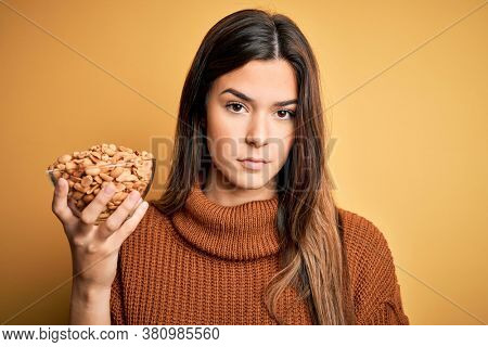 Young beautiful girl holding bowl of salty peanuts standing over yellow background with a confident expression on smart face thinking serious