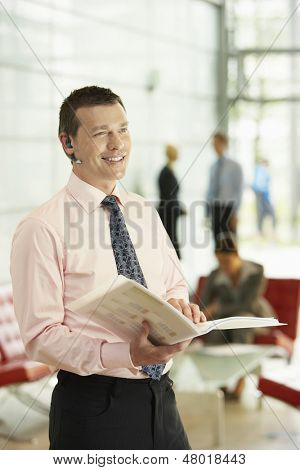 Happy middle aged businessman using Bluetooth while holding folder in office with colleagues working in background
