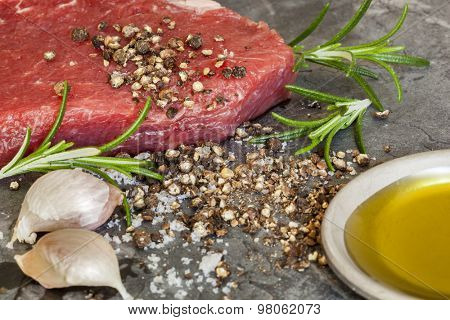 Raw beef steak with peppercorns, rosemary, garlic cloves and olive oil.